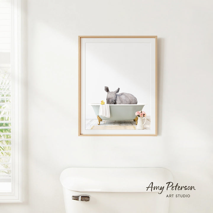 A framed image of a rhinoceros sitting in a bathtub, with a vase of flowers and a white toilet in the foreground.