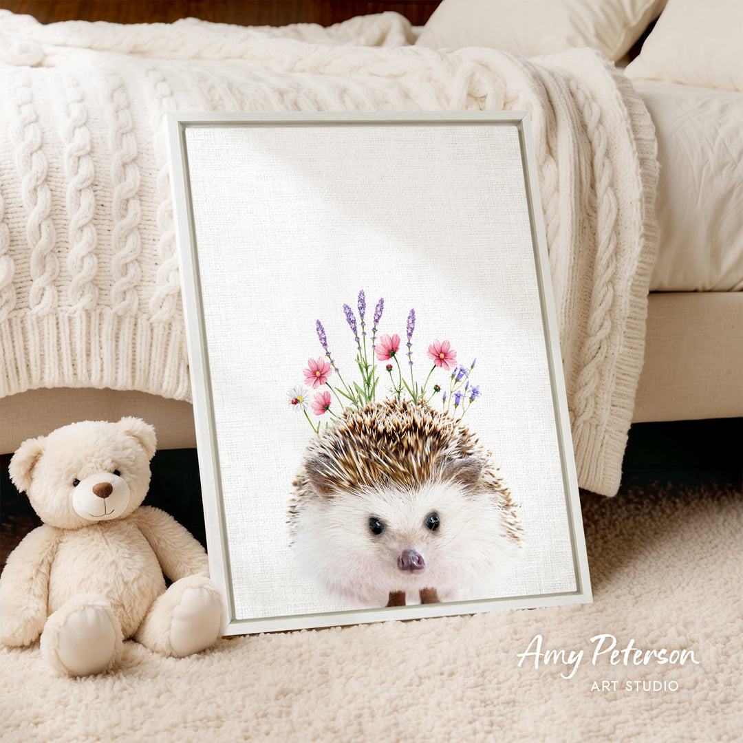 A framed print of a hedgehog with flowers is displayed on a bed, accompanied by a teddy bear.