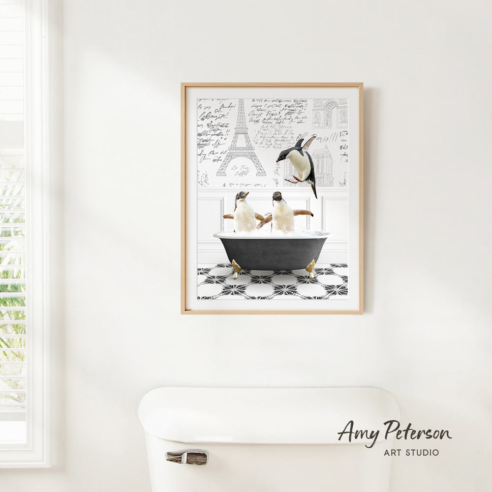 A framed print of two penguins sitting in a bathtub is hanging on a white wall, with a window and a toilet visible in the background.