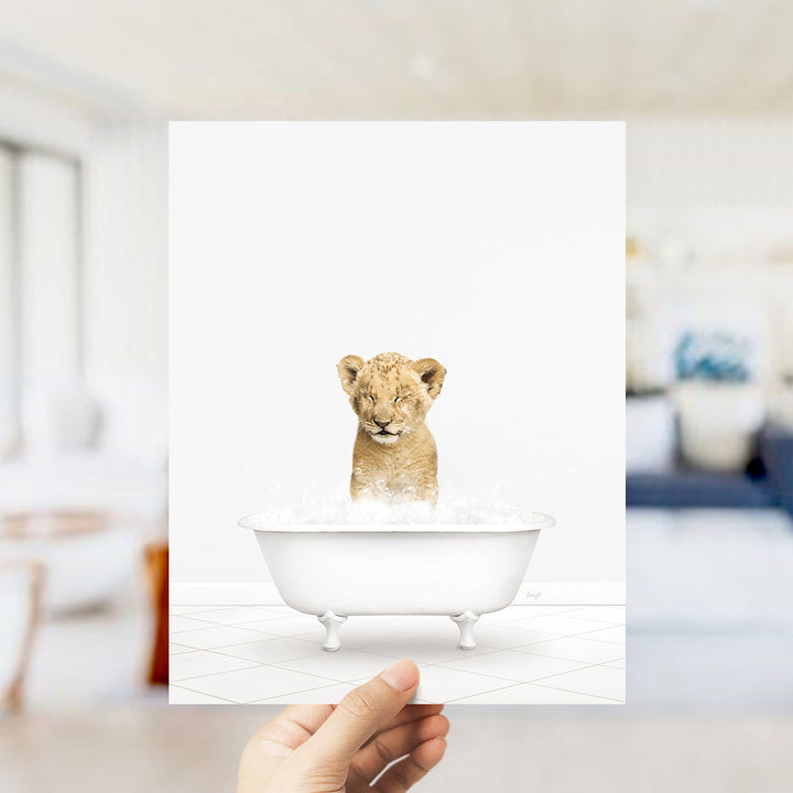 A hand holds up a framed image of a young lion cub sitting in a bathtub filled with water.