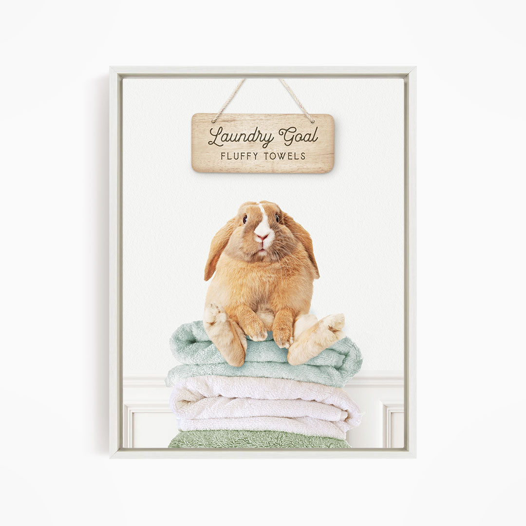 A fluffy rabbit sitting on a stack of towels, with a wooden sign above that says "Laundry Goal Fluffy Towels".