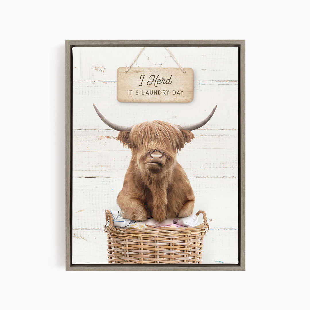 A framed image of a Highland cow sitting in a wicker basket, with a sign above it that says "It's laundry day".