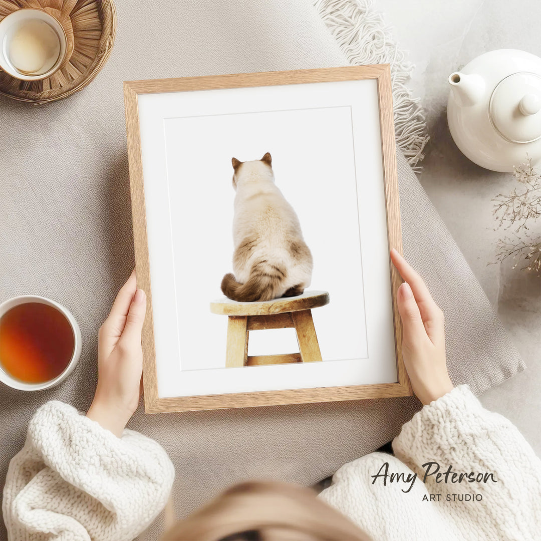 A person is holding a framed picture of a cat sitting on a wooden stool, with a teapot and a cup of tea nearby.