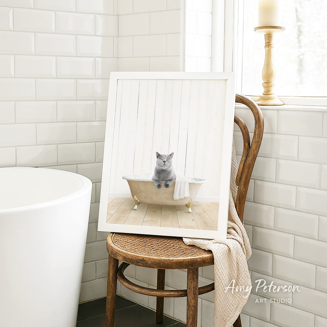 A framed image of a gray cat sitting in a bathtub, with a towel draped over the side of the tub, is displayed on a wooden chair in a bathroom setting.