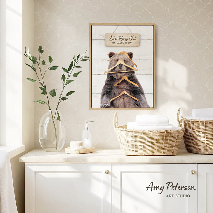 a framed picture of a bear with a sign above it that says "Let's Hang Out on Laundry Day". The bear is holding three clothes hangers in its mouth. The setting appears to be a bathroom, with a wicker basket and a vase of leaves on the counter.