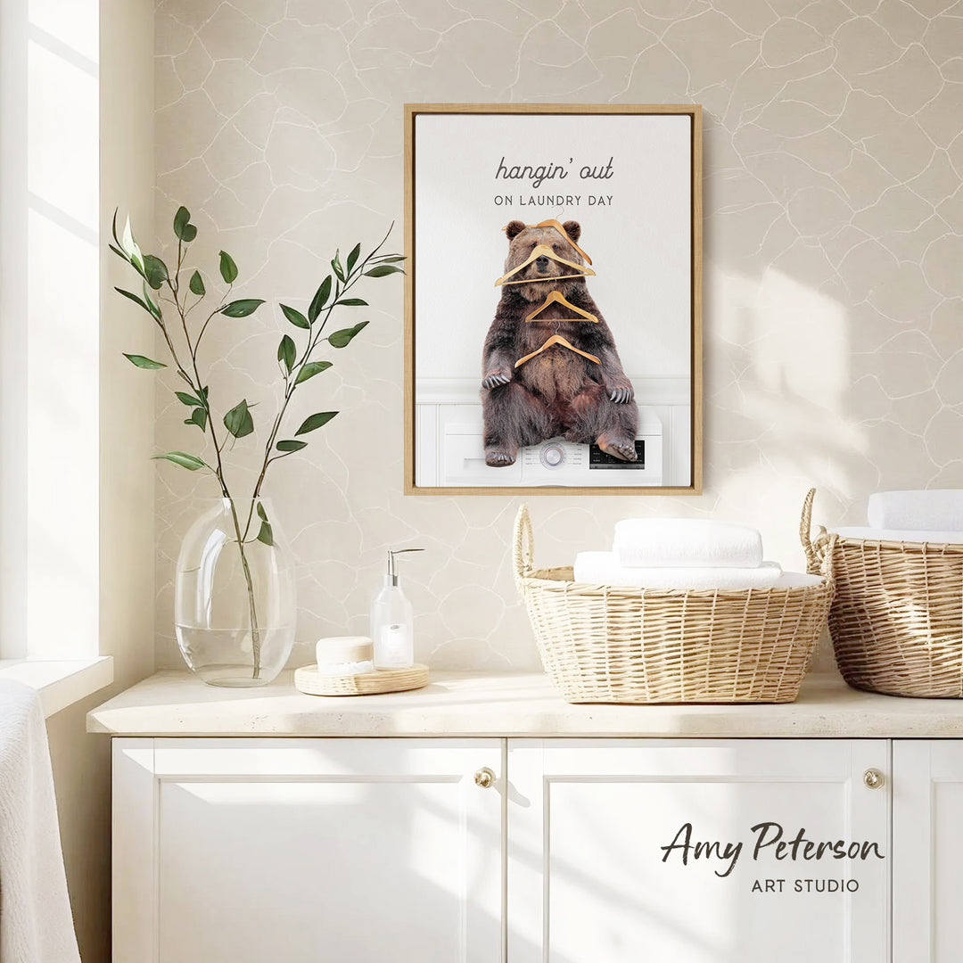 a framed poster of a bear sitting on a shelf, with the text "hangin' out on laundry day" written above it. The poster is displayed on a wall above a white cabinet, which has a wicker basket and a soap dispenser on it. There is also a plant in a clear vase on the cabinet.