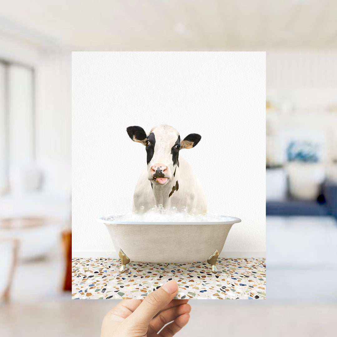 A hand holds up a framed image of a cow sitting in a bathtub filled with water, creating a whimsical and humorous scene.