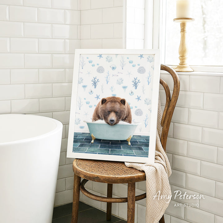 A framed image of a brown bear sitting in a bathtub, surrounded by blue and white floral patterns, is displayed on a wooden chair in a bathroom setting.
