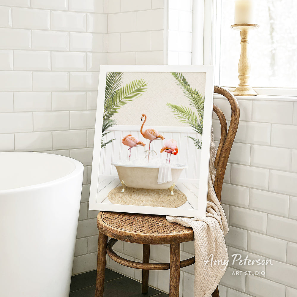 A framed image of three pink flamingos sitting in a white bathtub, surrounded by green plants, is displayed on a wooden chair in a bathroom setting.
