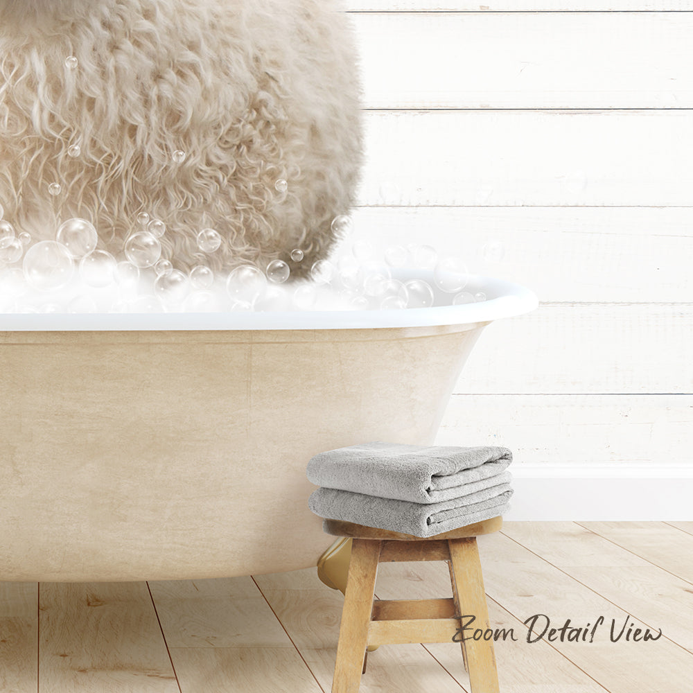 A fluffy white dog is standing in a bathtub filled with bubbles, with a towel and stool nearby.