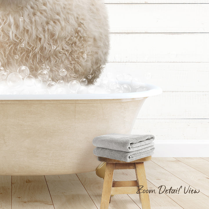 A fluffy white dog is standing in a bathtub filled with bubbles, with a towel and stool nearby.