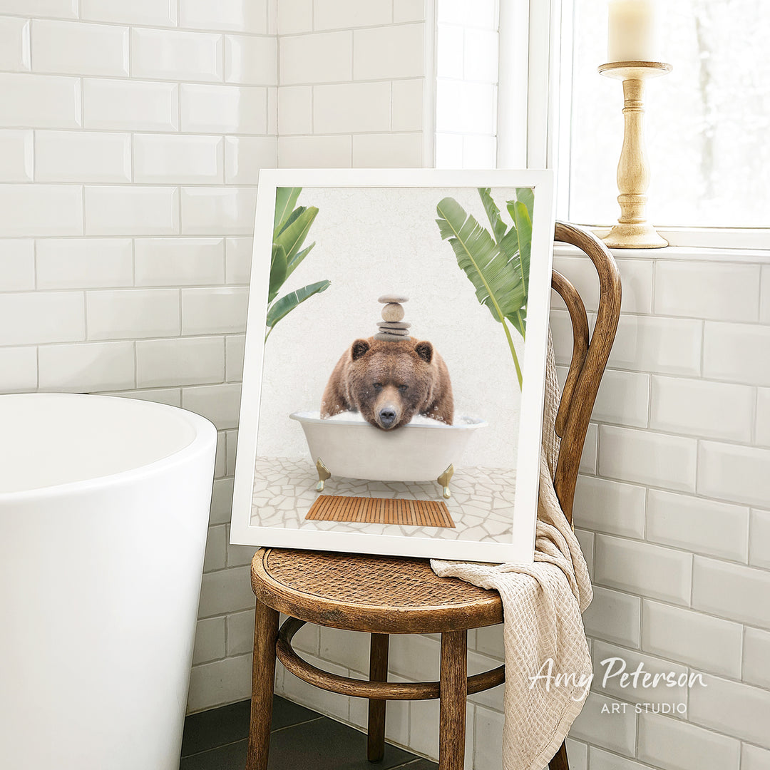 A framed image of a brown bear sitting in a bathtub, surrounded by tropical plants, is displayed on a wooden chair in a bathroom setting.