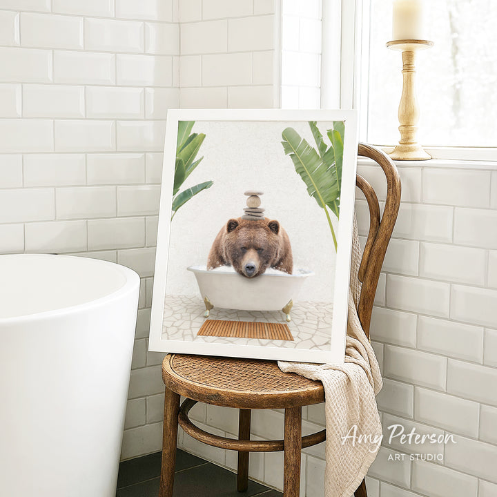 A framed image of a brown bear sitting in a bathtub, surrounded by tropical plants, is displayed on a wooden chair in a bathroom setting.