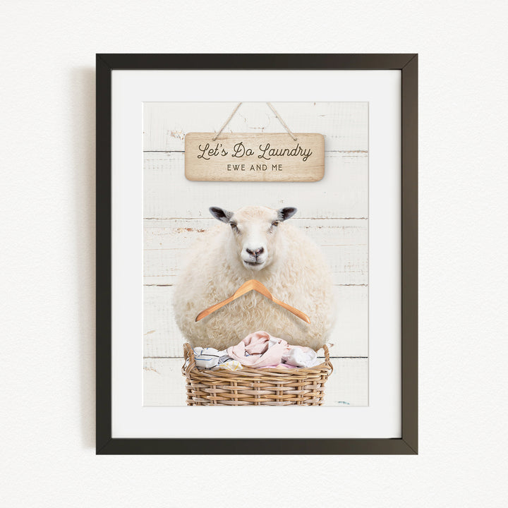 A framed image of a sheep sitting in a basket filled with laundry, with a sign above it that says "Let's Do Laundry EWS AND ME".