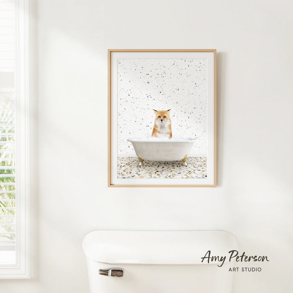 A framed image of a cat sitting in a bathtub, surrounded by a speckled background, is hanging on a white wall in a bathroom.