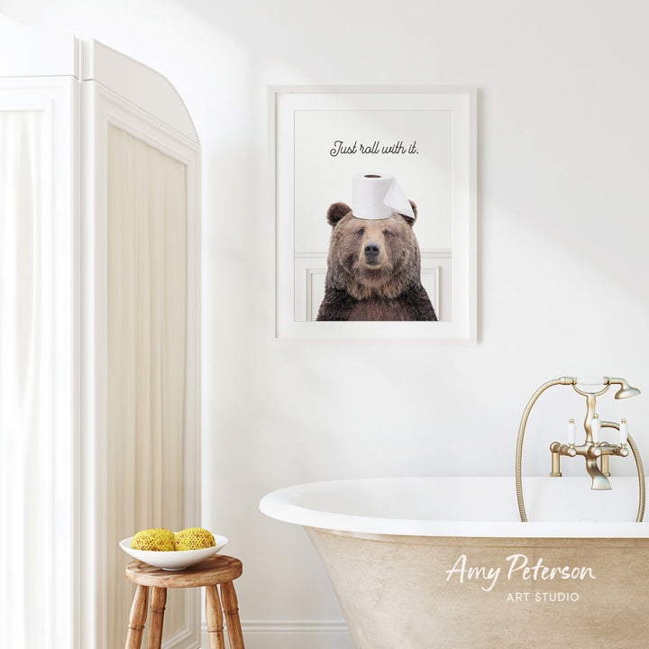 a bathroom with a framed picture of a bear wearing a toilet paper roll on its head, sitting on a stool next to a bowl of fruit.