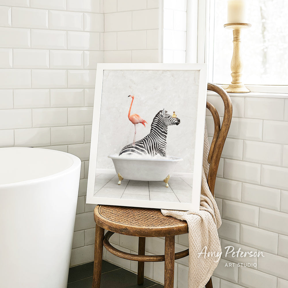 A framed painting of a zebra sitting in a bathtub, with a pink flamingo perched on its back, is displayed on a wooden chair in a bathroom setting.