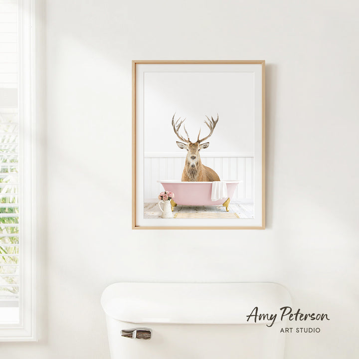 A framed image of a deer sitting in a pink bathtub, with a vase of flowers and a white toilet in the foreground.