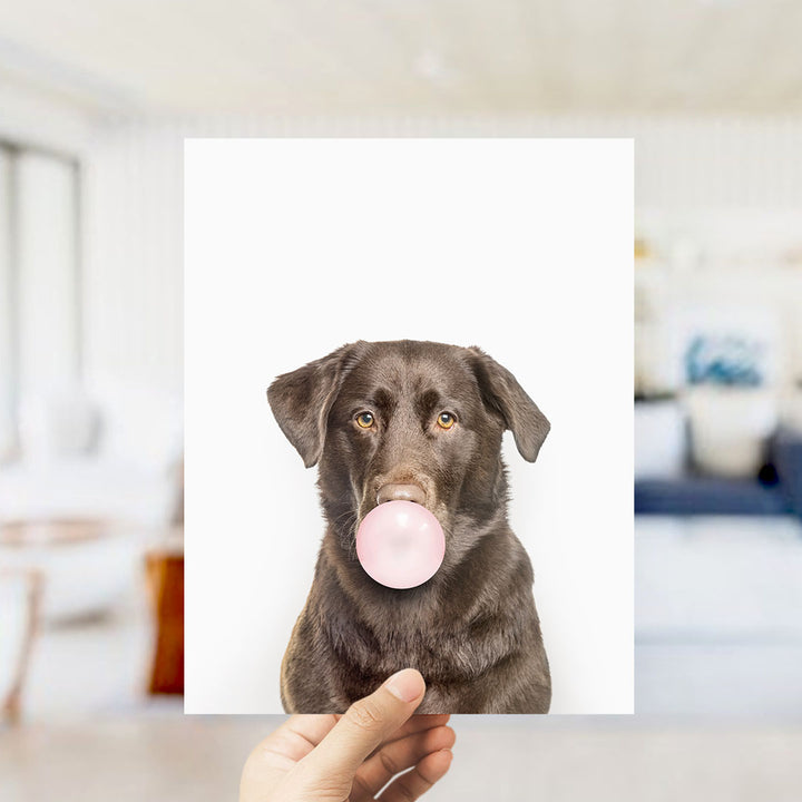 A hand holds up a photograph of a brown dog blowing a bubble with a pink bubble gum bubble.