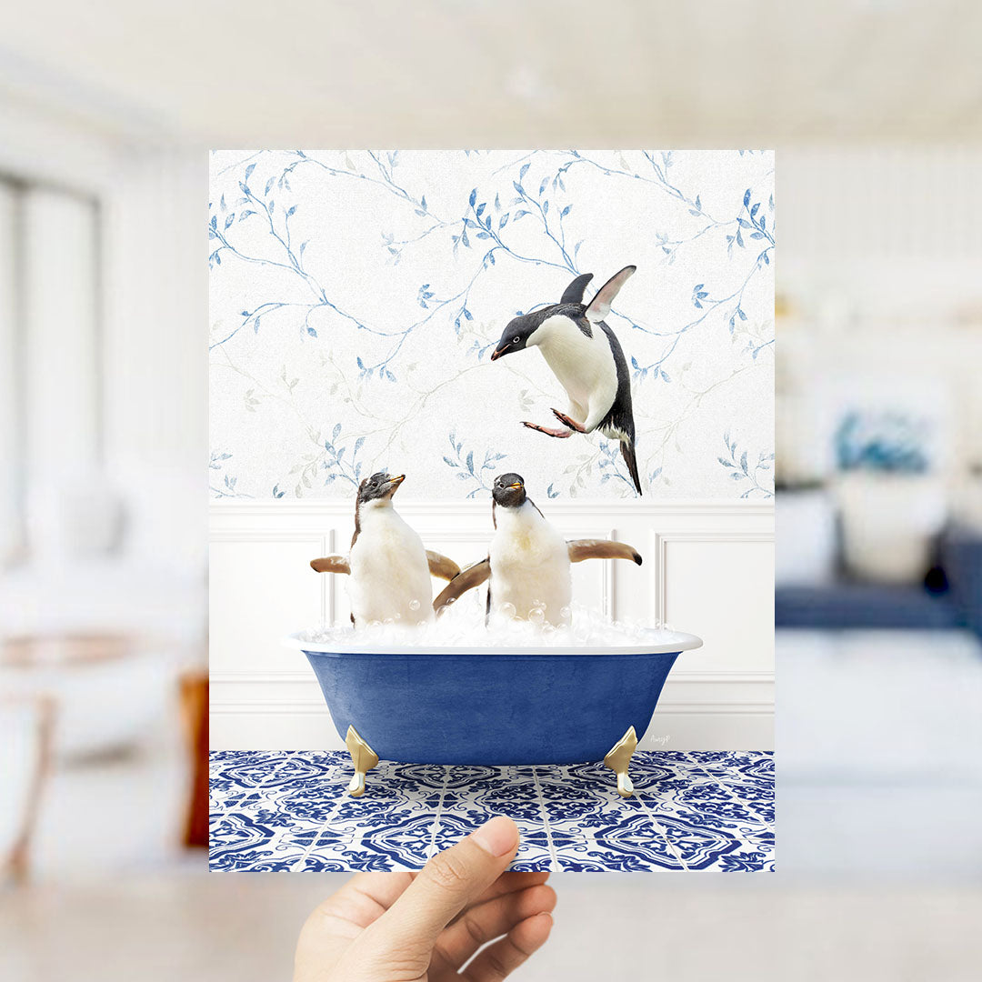 A hand holds up a framed photograph of two penguins sitting in a blue bathtub, with one penguin flying towards the other.