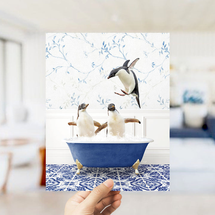A hand holds up a framed photograph of two penguins sitting in a blue bathtub, with one penguin flying towards the other.