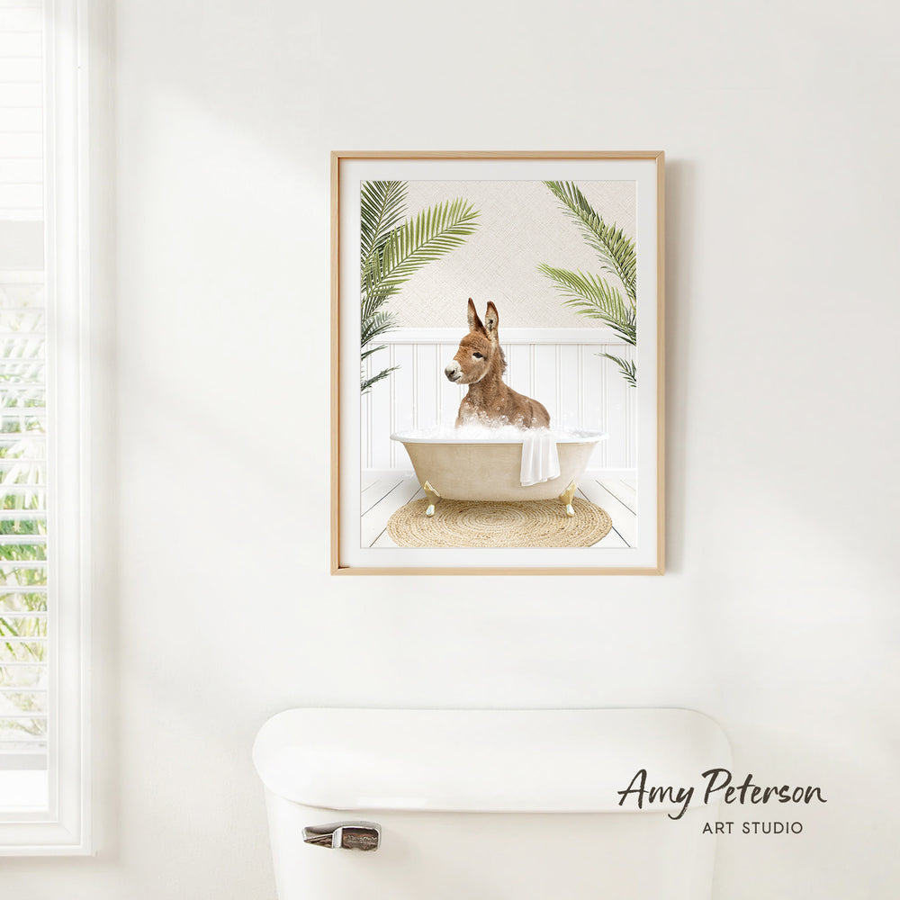 A framed print of a donkey sitting in a bathtub, surrounded by palm leaves, is hanging on a white wall in a bathroom.