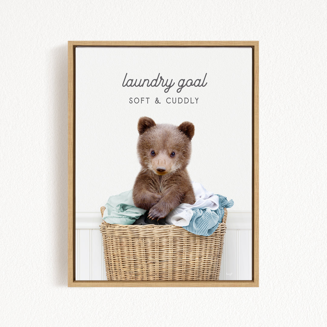 A cute brown bear sitting in a wicker laundry basket filled with clothes, with the text "laundry goal SOFT & Cuddly" displayed above the bear.