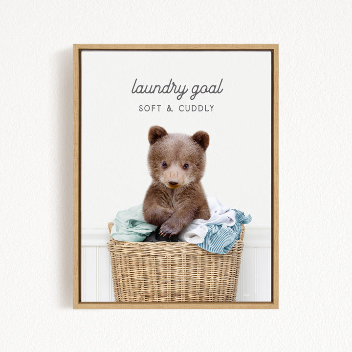 A cute brown bear sitting in a wicker laundry basket filled with clothes, with the text "laundry goal SOFT & Cuddly" displayed above the bear.