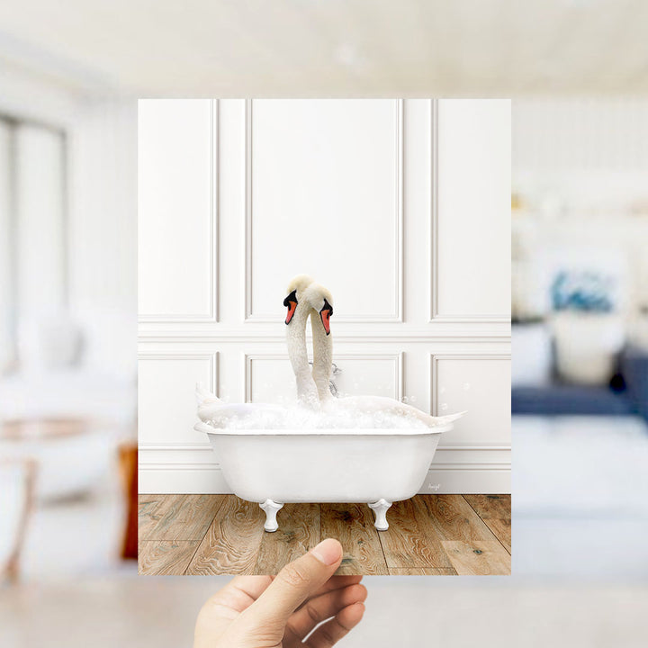 A hand holds up a photograph of a white swan sitting in a bathtub filled with bubbles, with a blurred background suggesting a bathroom setting.