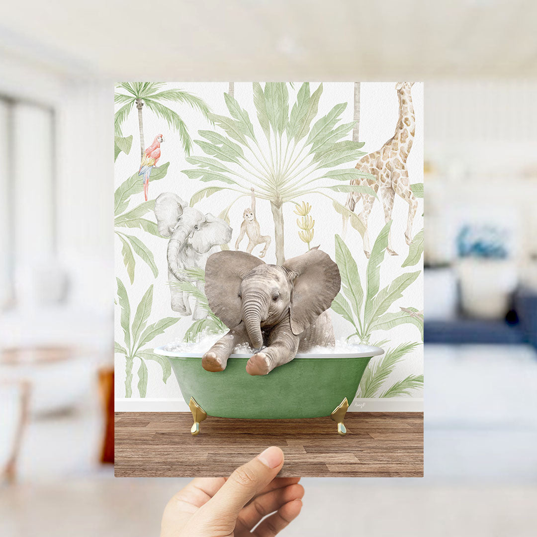A hand holds up a framed print featuring a whimsical scene of an elephant sitting in a green bathtub, surrounded by tropical foliage and other animals.