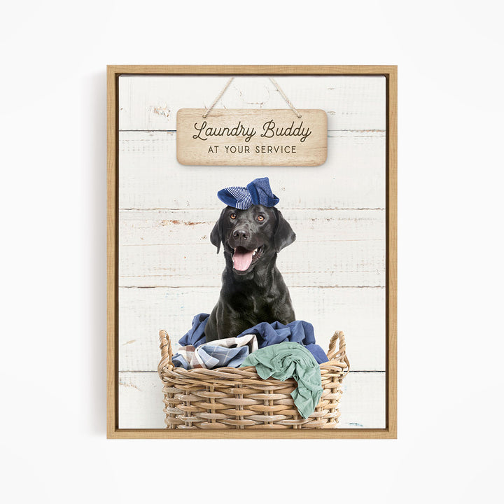 A black dog wearing a blue hat sits in a wicker basket filled with clothes, with a wooden sign above that reads "Laundry Buddy at your service".