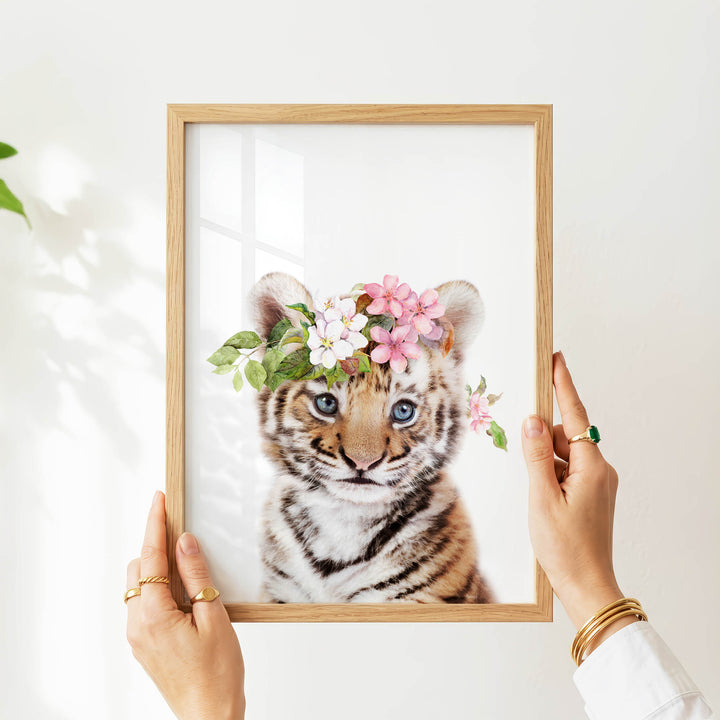 A person's hands are holding a picture frame with a young tiger cub inside it, wearing a floral crown.