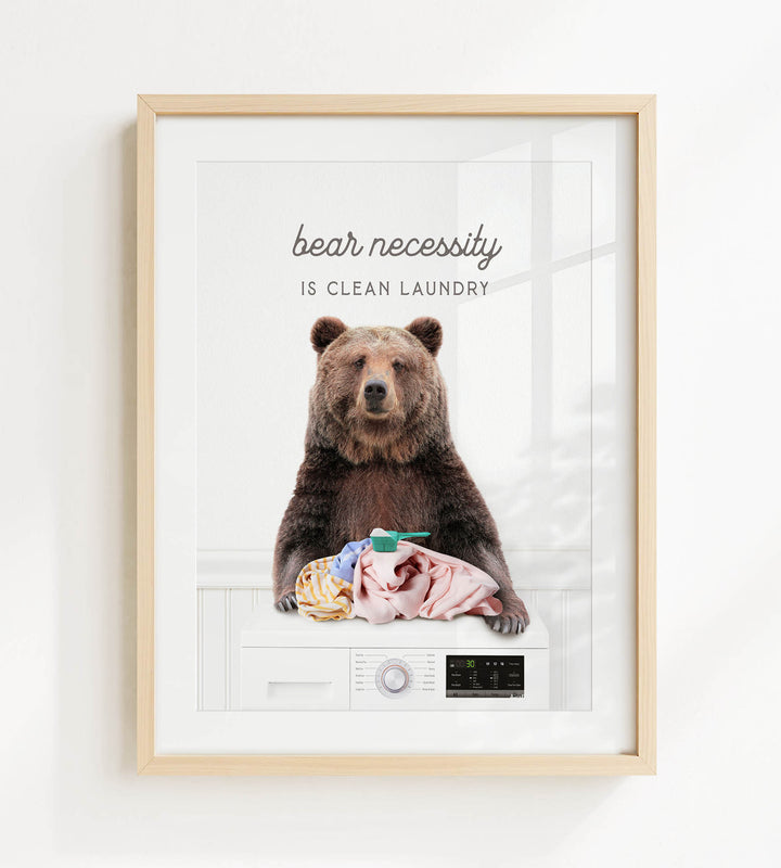 A framed image of a brown bear sitting on a washing machine, with the text "bear necessity is clean laundry" above it.
