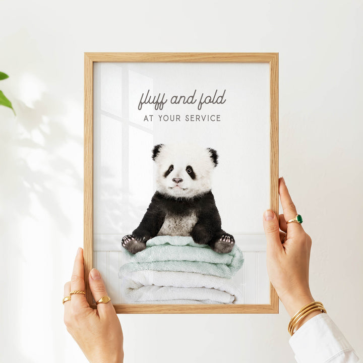 A person's hands are holding a framed picture of a panda bear sitting on a stack of towels, with the text "fluff and fold at your service" written above the image.