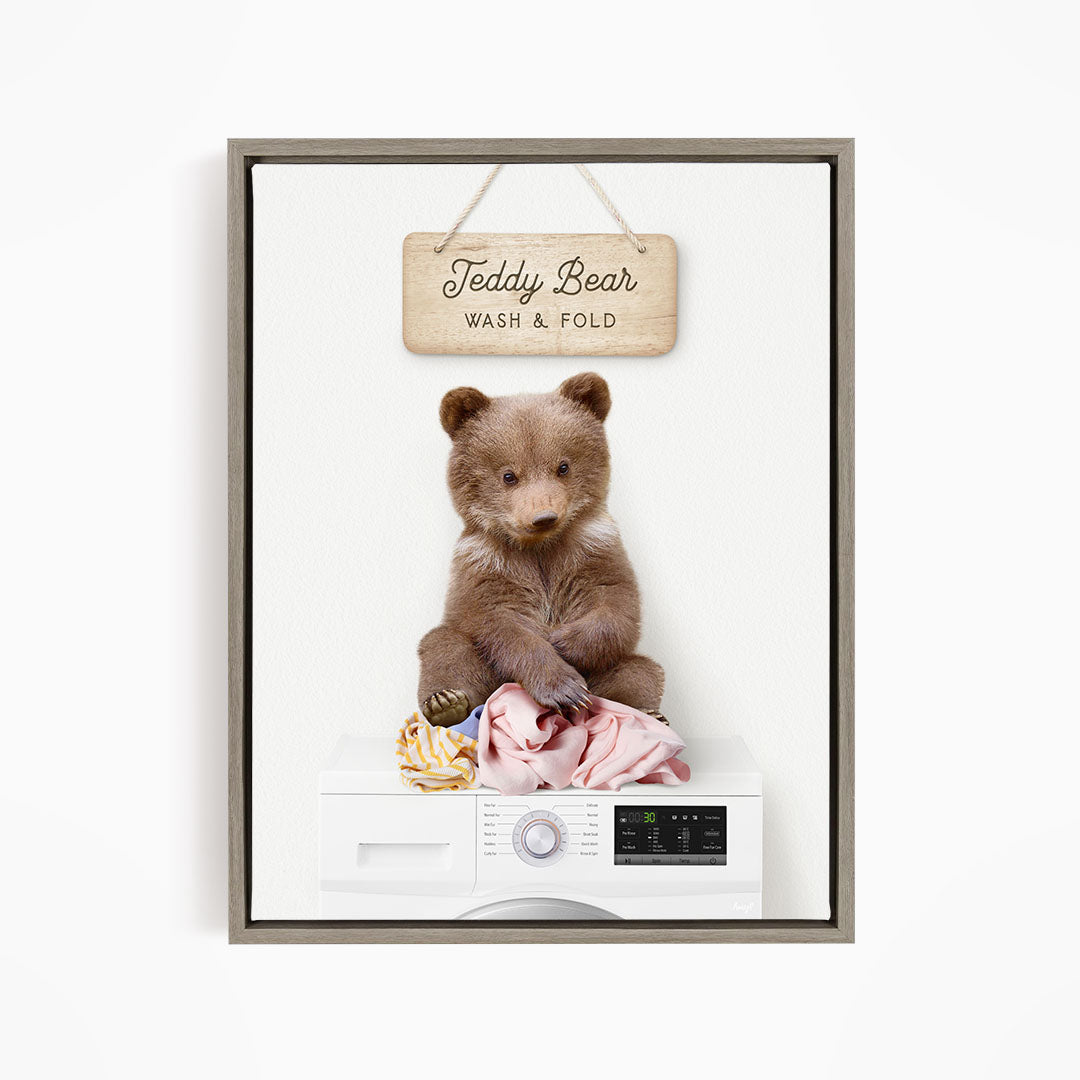A teddy bear sitting on a pile of laundry next to a washing machine, with a sign above that says "Teddy Bear Wash & Fold".