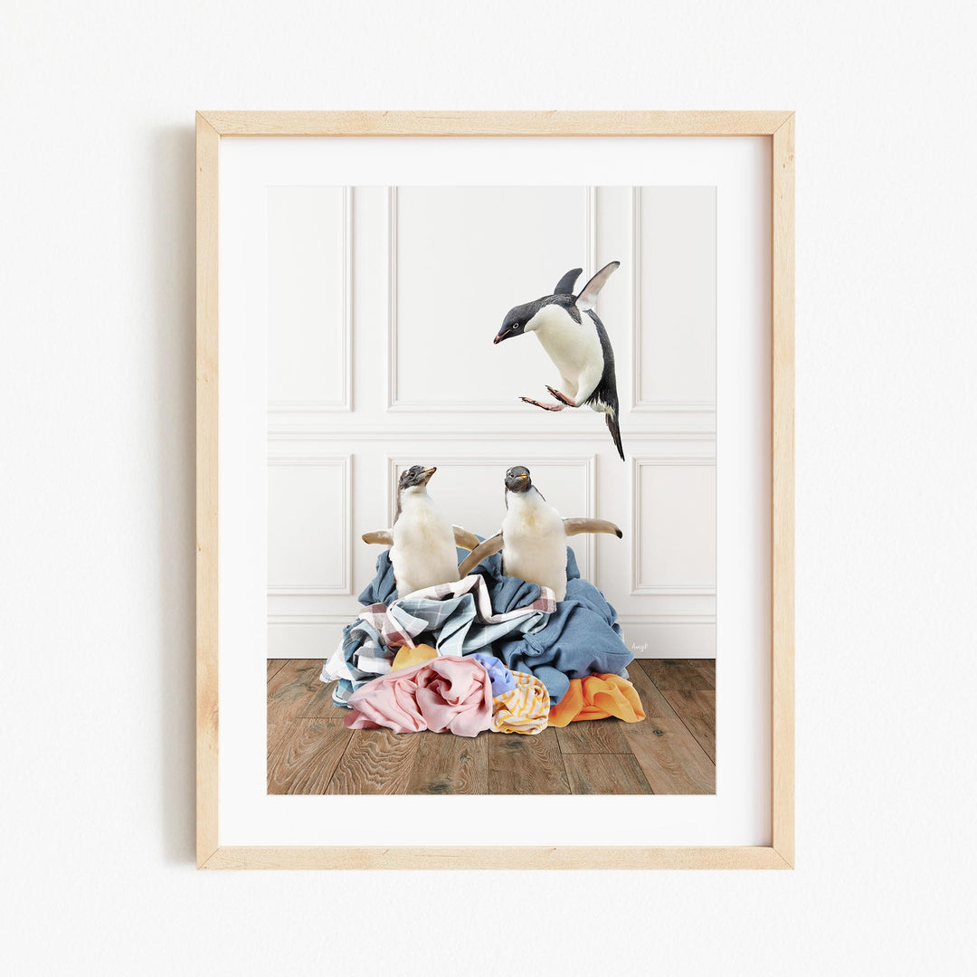 A framed photograph of two penguins standing on a pile of clothes, with one penguin flying in the air above the other.