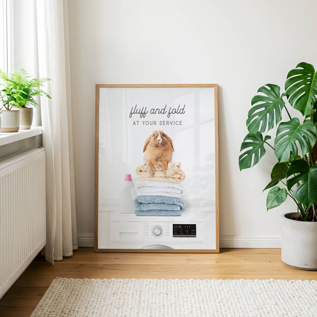 A framed poster with a rabbit sitting on a stack of folded towels, accompanied by the text "fluff and fold at your service" in a white frame.