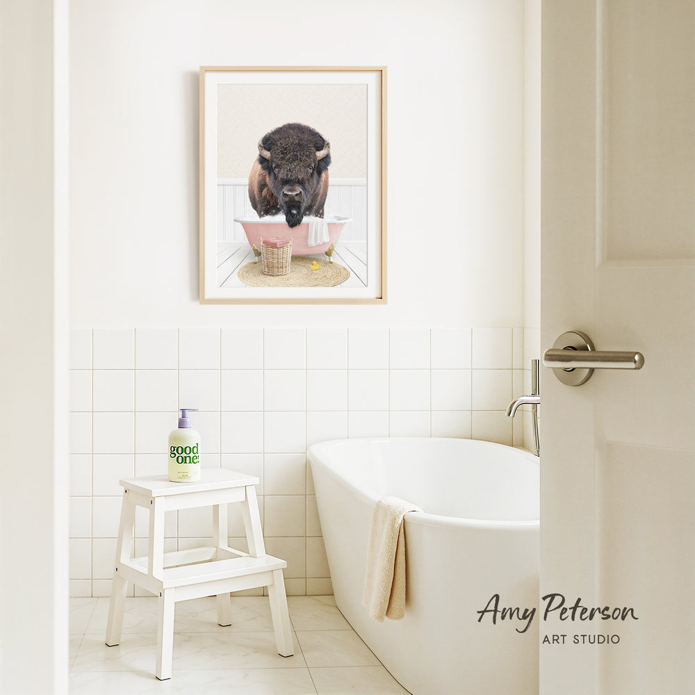a bathroom with a framed picture of a buffalo sitting in a bathtub, surrounded by a pink towel and a white stool.