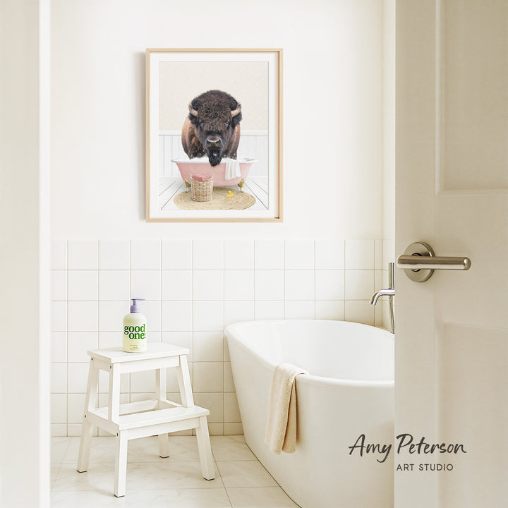 a bathroom with a framed picture of a buffalo sitting in a bathtub, surrounded by a pink towel and a white stool.