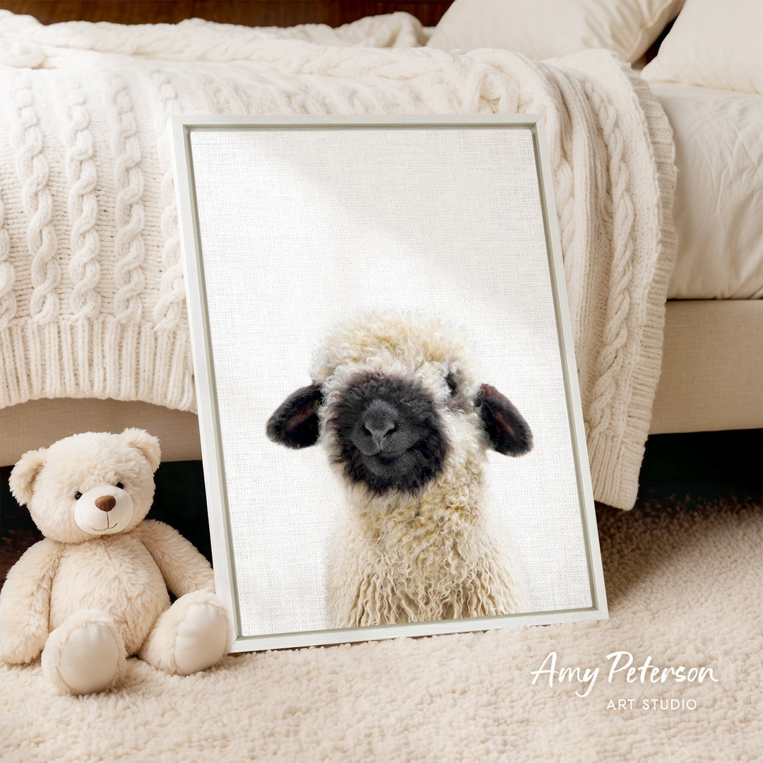 A framed image of a sheep with a black face is displayed on a white wall, accompanied by a teddy bear sitting next to it.