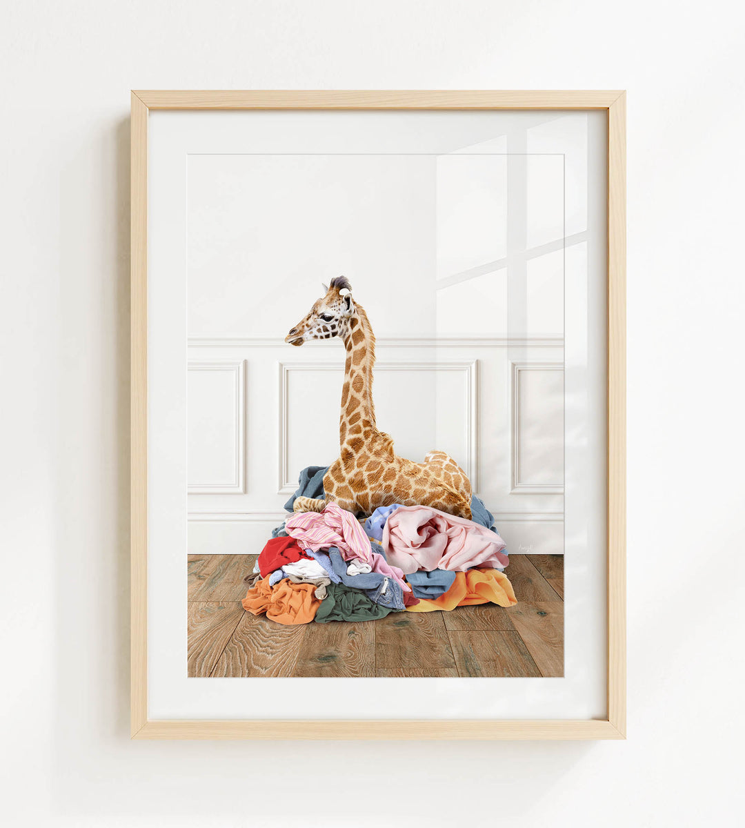 A giraffe is sitting on a pile of clothes on a wooden floor, with a white wall in the background.