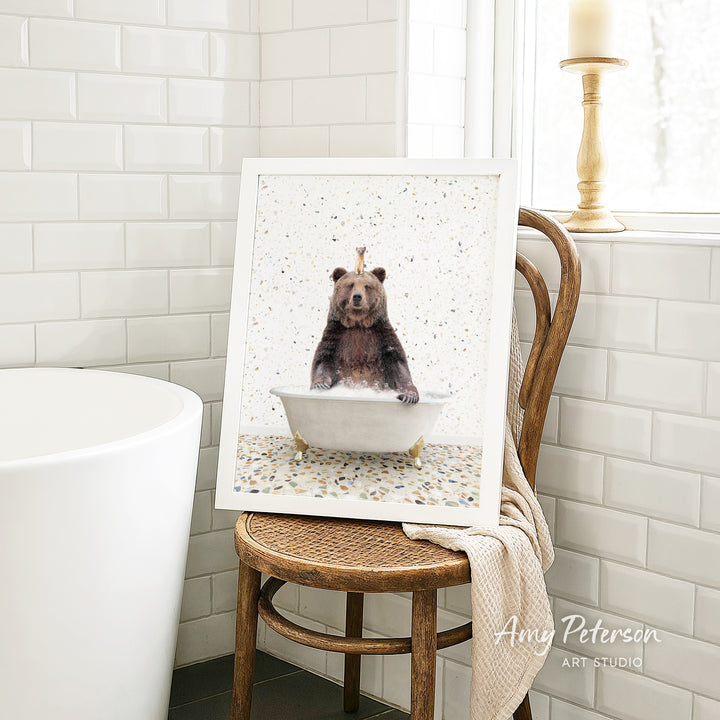 A framed image of a bear sitting in a bathtub, with a towel draped over the back of a chair in a bathroom setting.