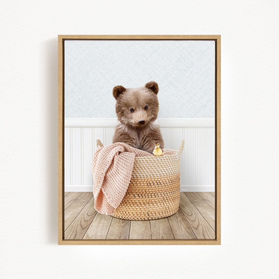 A brown bear sitting in a woven basket, with a pink blanket and a small yellow toy duck nearby.