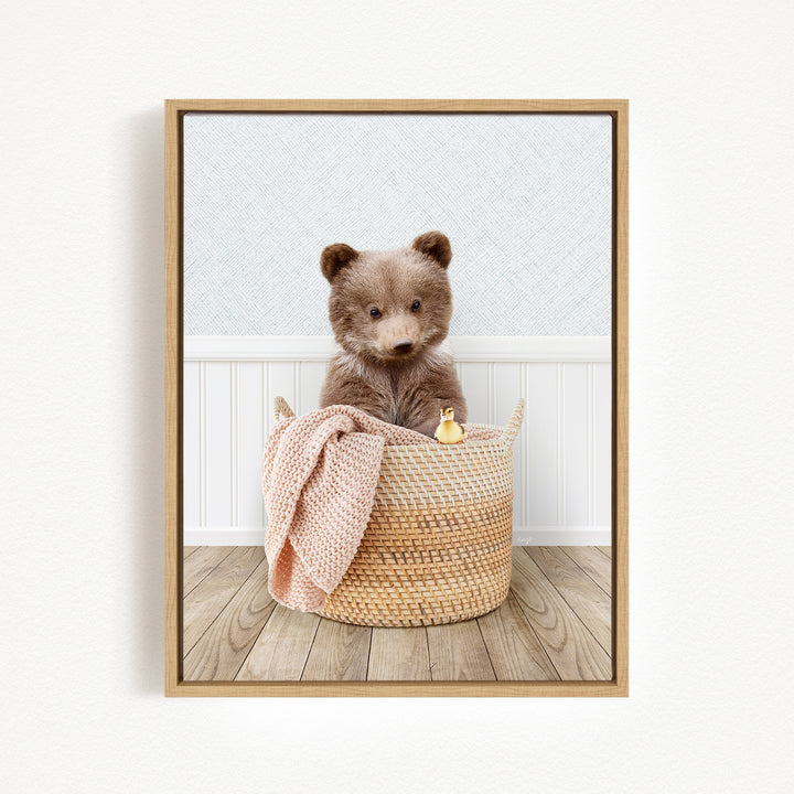 A brown bear sitting in a woven basket, with a pink blanket and a small yellow toy duck nearby.