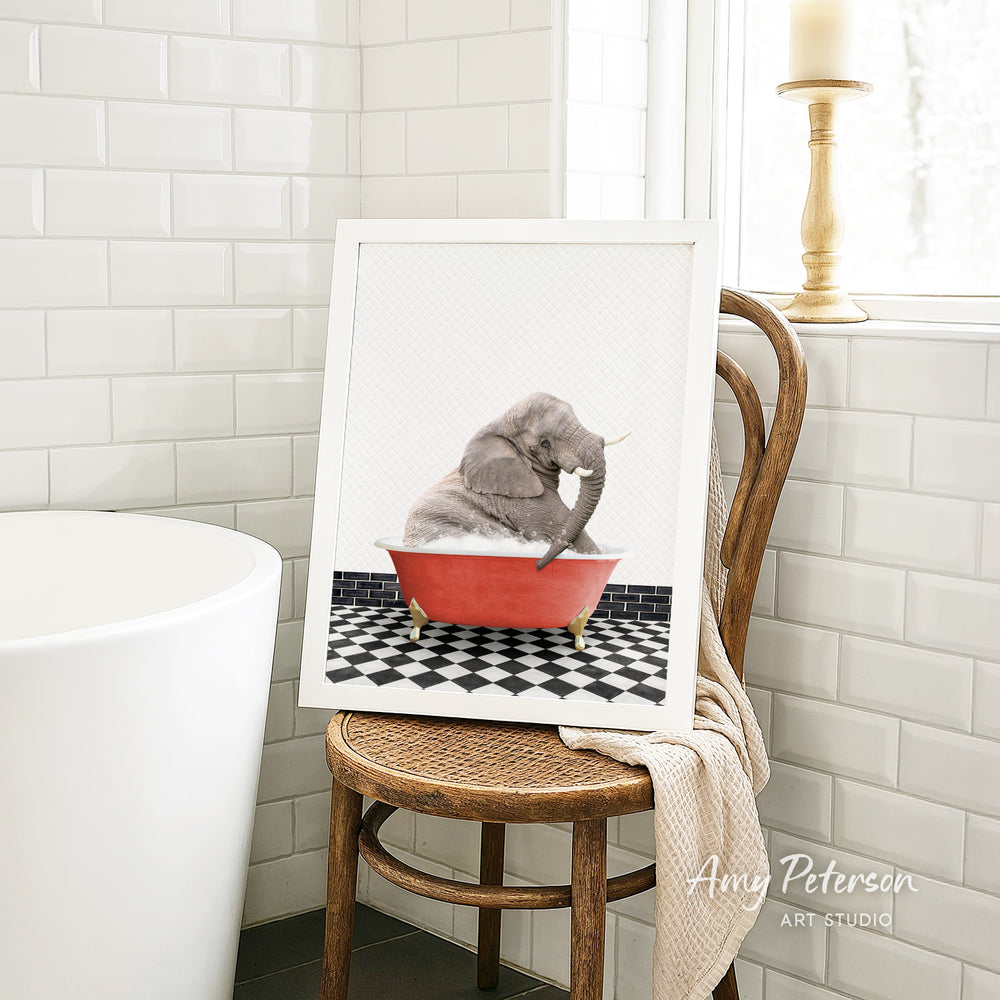 A framed image of a gray elephant sitting in a red bathtub, surrounded by a black and white checkered floor, is displayed on a wooden chair in a bathroom setting.
