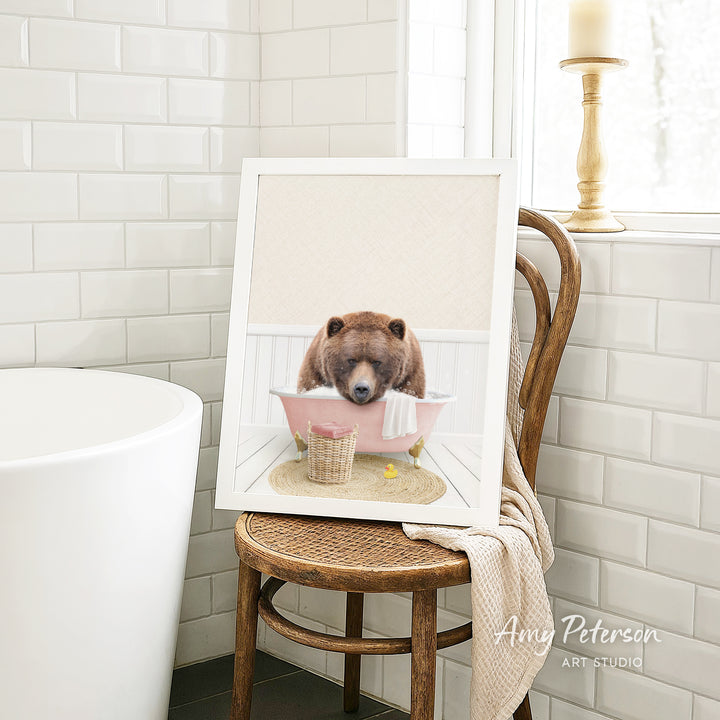A framed image of a brown bear sitting in a pink bathtub, with a towel and a basket nearby, is displayed on a wooden chair in a bathroom setting.