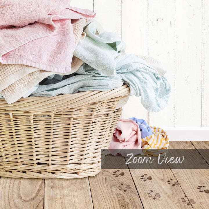 A wicker basket filled with various folded clothes sits on a wooden floor, with paw prints visible on the floor.