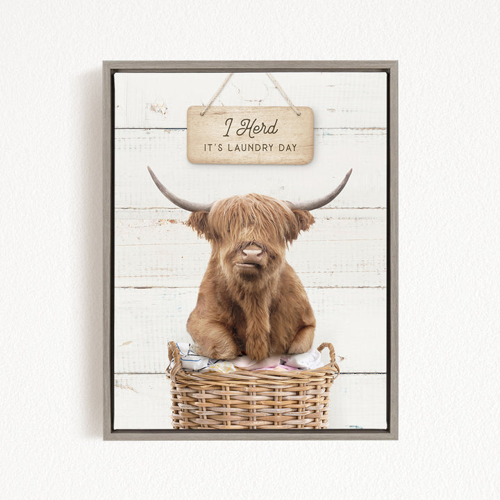 A framed image of a Highland cow sitting in a wicker basket, with a sign above it that says "It's laundry day".