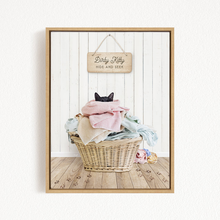 A framed image of a cat sitting in a basket filled with clothes, with a sign above the basket that reads "Dirty Kitty HIDE AND SEEK".