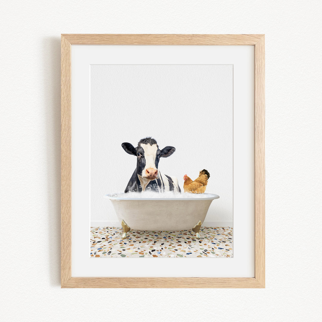 A black and white cow is sitting in a white bathtub, with a chicken standing nearby, in a framed image.